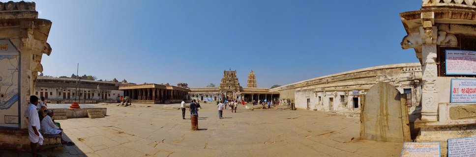 Wide view of Virupaksha Temple complex showing ornate gopuram towers, stone courtyard with visitors, and colonnaded corridors under blue sky in Hampi, India