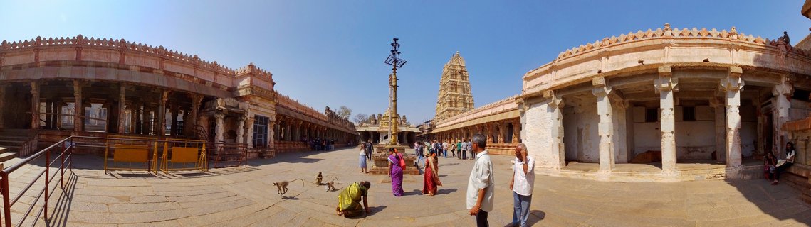 Wide view of Virupaksha Temple courtyard in Hampi showing stone colonnaded corridors on both sides, central lamp pillar, temple gopuram in background, visitors in colorful saris and traditional clothing, monkeys on stone pavement, clear blue sky