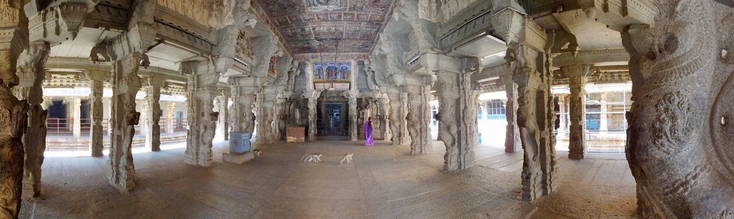 Interior view of historic Hindu temple showing rows of ornately carved stone pillars with sculptures, painted ceiling, and stone floor