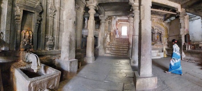 Interior of ancient stone temple showing multiple carved granite pillars, stone floor, stairs leading to bright doorway, woman in blue sari walking, and traditional water fountain feature