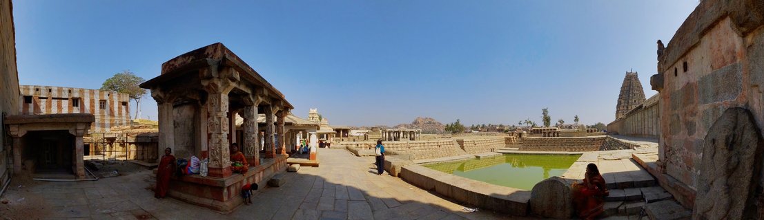 Wide view of ancient stone temple courtyard in Hampi India showing carved pillared pavilion, rectangular sacred tank with green water, temple tower in distance, and people in traditional clothing under clear blue sky