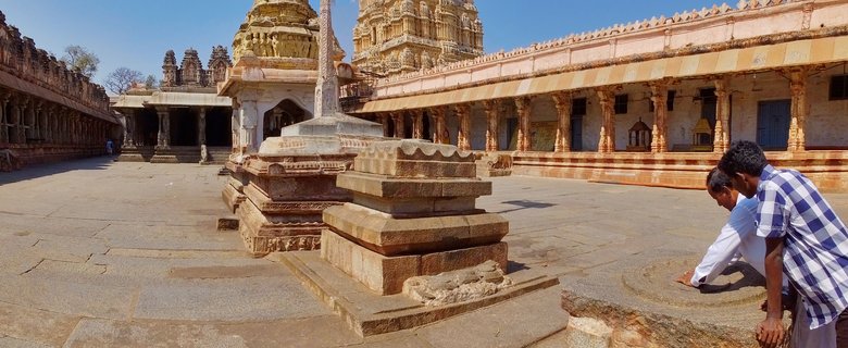Stone temple courtyard in Hampi, India showing ancient Dravidian architecture with carved pillars, temple towers, and visitors exploring the historic complex under clear blue sky