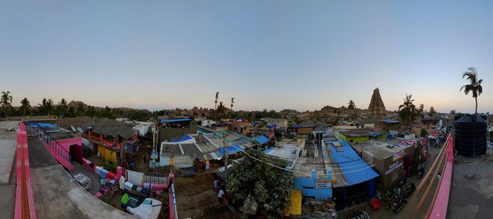 Panoramic aerial view of Hampi village at twilight showing colorful building rooftops, palm trees, and a Hindu temple tower against a blue gradient sky with rocky hills in the distance