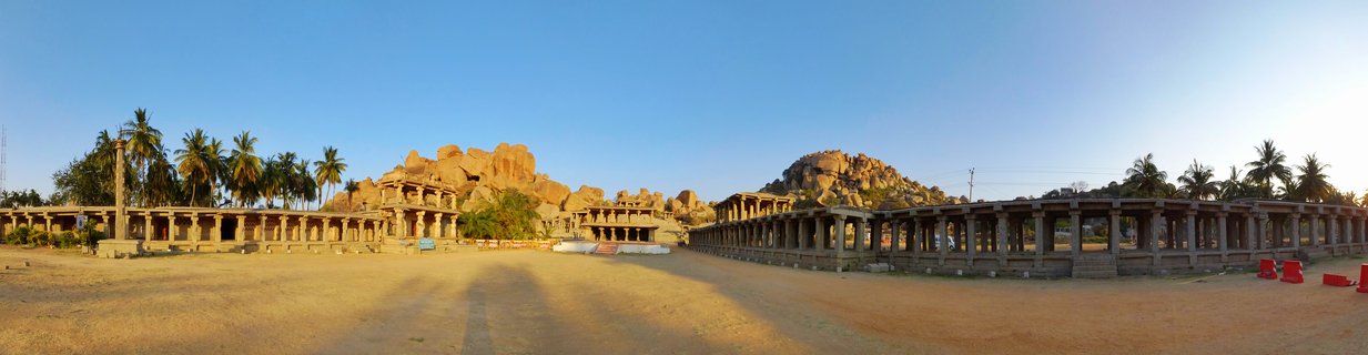 Ancient stone-pillared temple halls at Vittala Temple complex in Hampi with large boulder rock formations and palm trees under clear blue sky