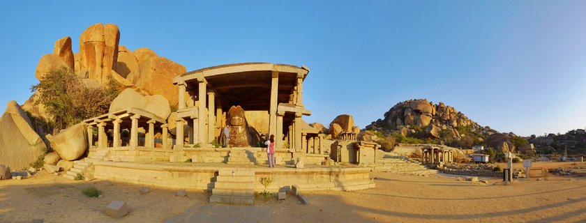 Ancient stone temple ruins with carved pillars and pavilion structure surrounded by large boulder rock formations under clear blue sky at Hampi heritage site in India