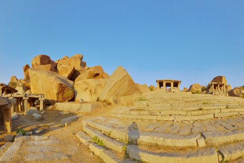 Ancient stone temple ruins with large granite boulders and pillared pavilions on terraced steps under clear blue sky at Hampi, India