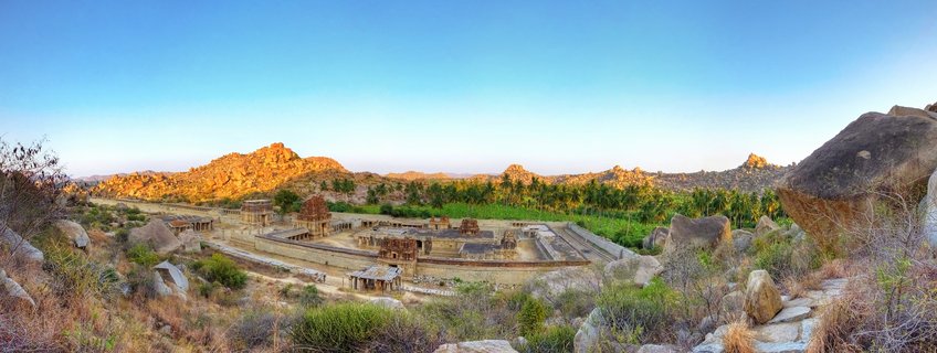 Panoramic view of ancient stone temple ruins surrounded by large boulders and rocky hills under clear blue sky at sunset in Hampi, India