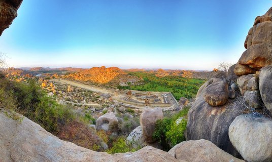Panoramic view of ancient stone temple ruins surrounded by large boulders and rocky hills in Hampi, India, with golden sunlight and clear blue sky