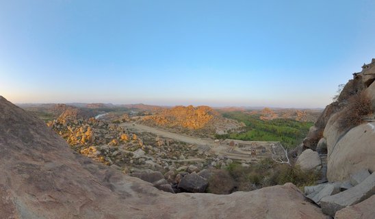 Panoramic view of ancient temple ruins among massive boulder formations in Hampi, India, with golden sunlight illuminating rocky hills and green vegetation in the valley below