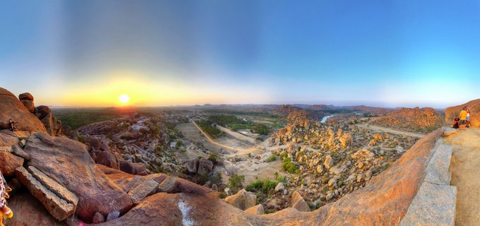 Panoramic sunset landscape showing Hampi's granite boulder fields in Karnataka, India, with golden light illuminating rocky terrain, scattered green vegetation, distant river, and clear blue-to-orange gradient sky