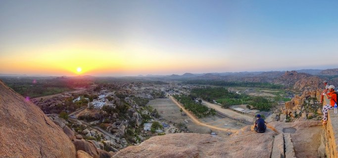 Panoramic sunset view over Hampi's ancient temple ruins and granite boulder landscape in Karnataka, India, with tourists sitting on rocky viewpoint in foreground