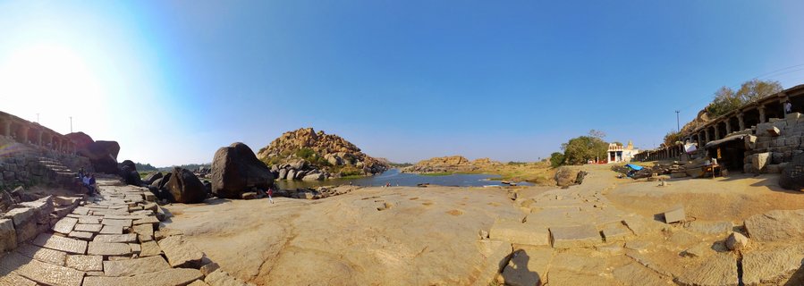 Panoramic photograph showing ancient stone ruins and large boulder formations along a river in Hampi, India, with clear blue sky and historic stone pathways in the foreground.