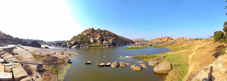 Wide river landscape showing traditional round coracle boats on the Tungabhadra River with rocky boulder hills and ancient stone ruins in Hampi, India under clear blue sky