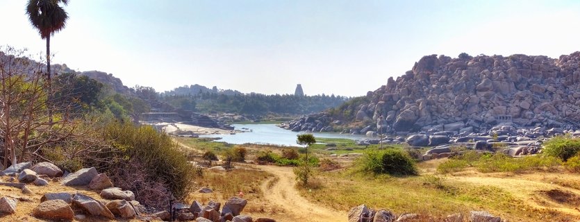 Panoramic landscape of Hampi showing rocky boulder-covered hills, Tungabhadra River, dirt path through dry grassland, palm tree, and Virupaksha Temple tower in the distance under clear sky