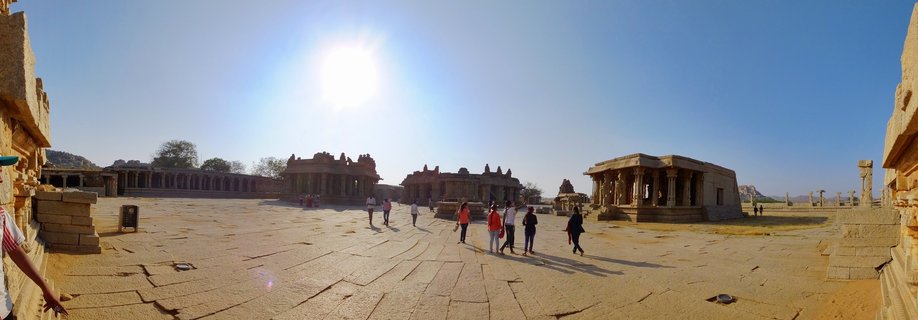 Wide view of ancient stone temple structures surrounding a large paved courtyard with visitors walking through the historic Hampi temple complex in India under bright sunlight