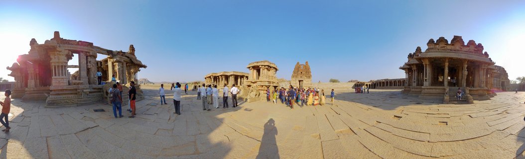 Ancient stone temple ruins with carved pillars and courtyards at Hampi with visitors walking among the historic structures under blue sky