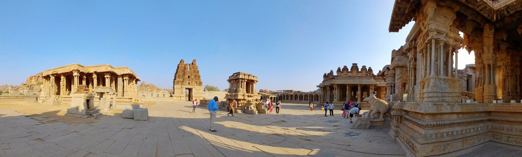 Ancient stone temple ruins with ornate carved pillars and mandapas in Hampi, India, with visitors walking on stone pavement under clear blue sky