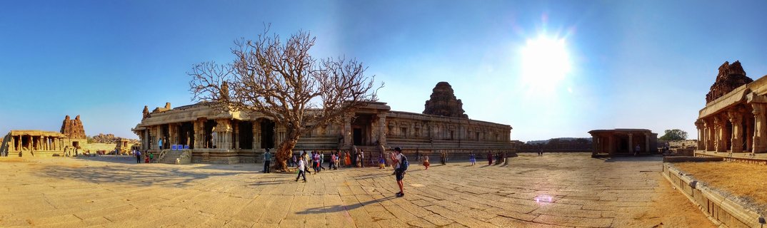 Panoramic view of ancient stone temple ruins in Hampi with tourists walking on paved courtyard, leafless tree, carved pillars, and bright sun in clear blue sky