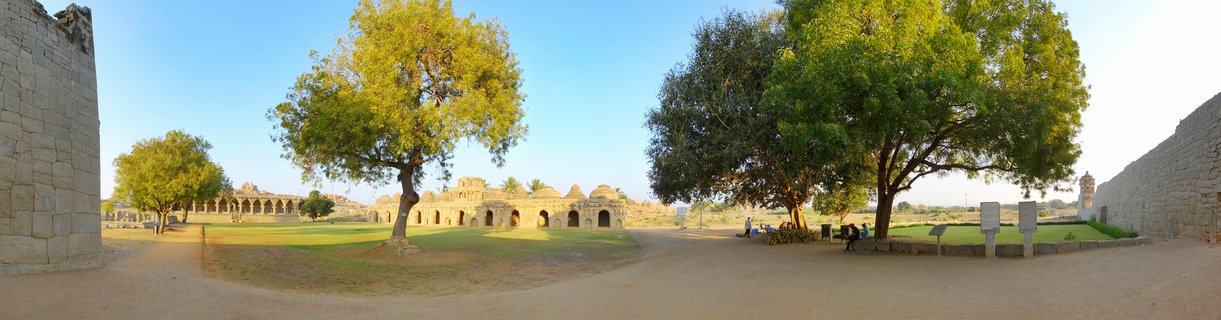 Panoramic view of ancient stone ruins at Hampi showing arched corridors, large green trees, and clear blue sky with visitors sitting in the courtyard