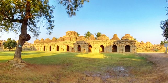 Historic stone building with eleven domed chambers and arched doorways at Hampi UNESCO site, with large tree and green lawn in foreground under clear blue sky