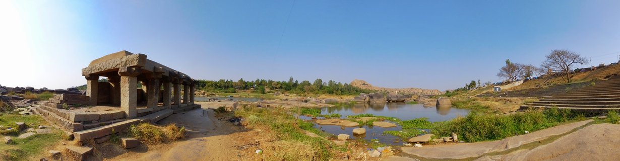 Ancient stone pavilion with carved pillars beside a river with boulders and vegetation at Hampi heritage site in India under clear blue sky