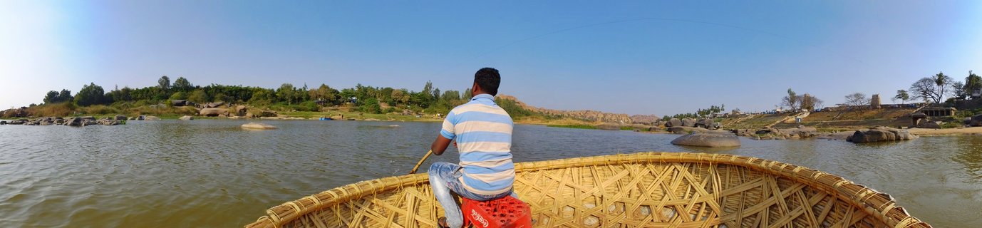 Person in blue and white striped shirt sitting in a round woven coracle boat on a river with rocky landscape and trees on the far bank under clear blue sky