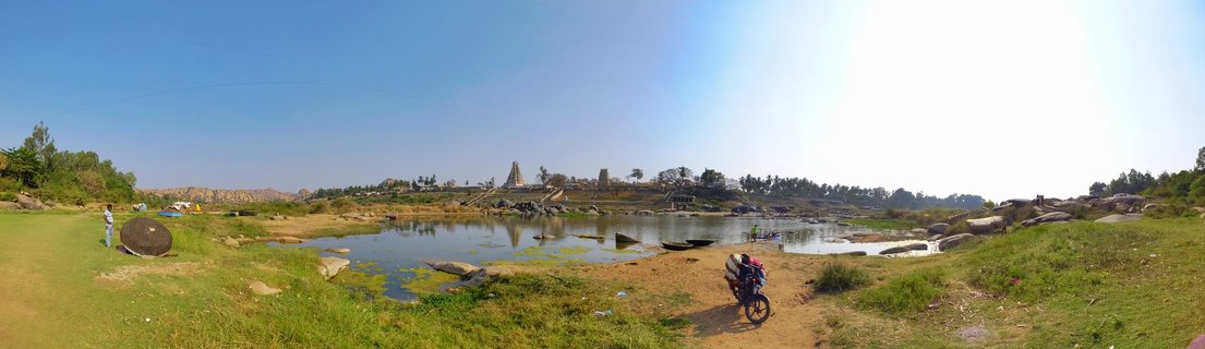 Panoramic landscape showing ancient Hindu temple towers in Hampi, India, with a river, rocky terrain, green grass, and a loaded motorcycle in the foreground under a clear blue sky
