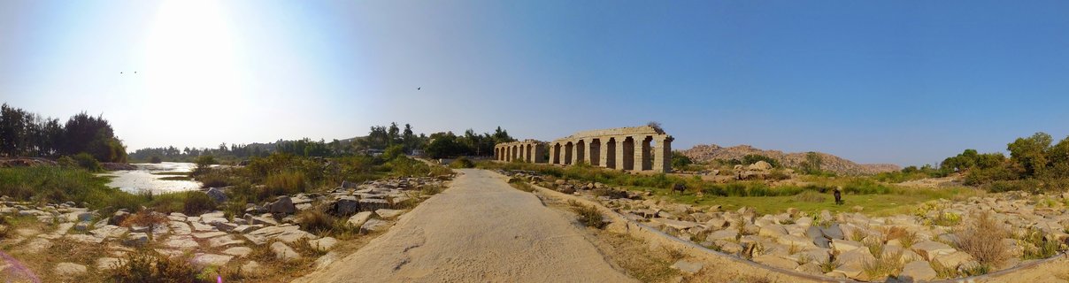 Ancient stone building with multiple arches along a rocky riverbank with dirt path leading toward it under clear blue sky in Hampi, India