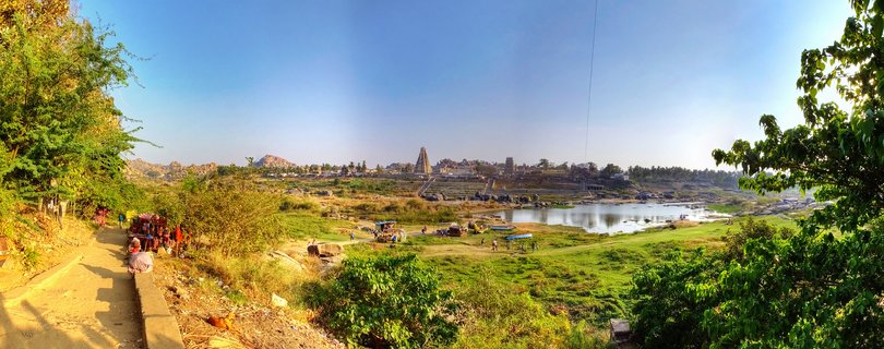 Panoramic view of Virupaksha Temple complex with tall tower visible across Tungabhadra River, green vegetation and trees in foreground, people on pathway at left, clear blue sky above Hampi, India