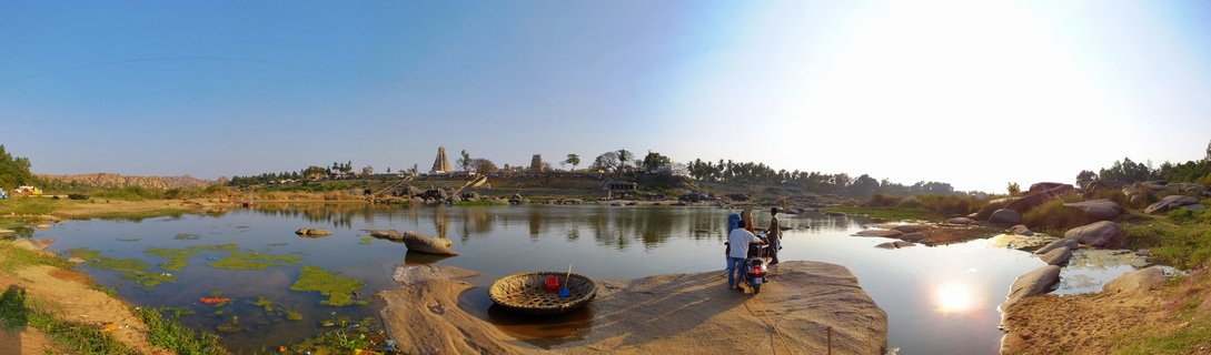 Wide panoramic landscape showing ancient Hindu temple tower across a calm river with green algae patches, traditional round coracle boat on rocky foreground, three people standing near motorcycle, large boulders scattered throughout, Hampi, India