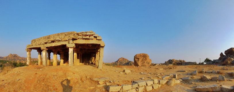 Ancient stone temple ruins with multiple carved pillars standing on dry earth with scattered rocks, blue sky above, rocky hills in background at Hampi, India