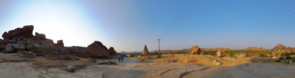 Wide landscape showing ancient stone temple tower and large boulder formations with two people walking on rocky ground under clear blue sky in Hampi, India