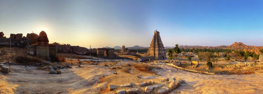 Ancient stone temple with tall gopuram tower at sunset in Hampi ruins, Karnataka, India, with rocky foreground and boulder-strewn landscape under clear blue sky