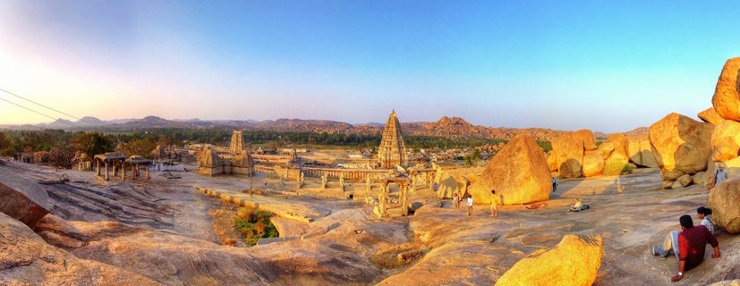 Ancient Virupaksha Temple gopuram tower rises above granite boulder formations and temple ruins during golden hour in Hampi, Karnataka, India, with tourists exploring the UNESCO World Heritage Site under clear blue sky