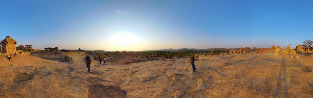 Panoramic view of ancient stone temple ruins on rocky terrain at sunset with visitors walking among the structures and distant hills visible on the horizon under a clear blue sky