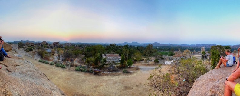 Panoramic sunset view from Hemakuta Hill showing tourists sitting on large granite boulders overlooking ancient temple ruins and rocky landscape of Hampi, India