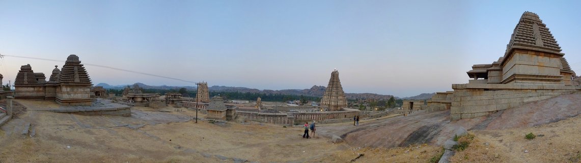 Ancient stone Hindu temple structures with pyramidal towers across rocky terrain in Hampi, India, with small figures of people walking in the foreground under clear evening sky