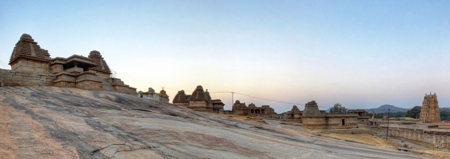 Panoramic view of ancient stone temples with pyramidal towers built on a large granite rock formation at sunset in Hampi, India