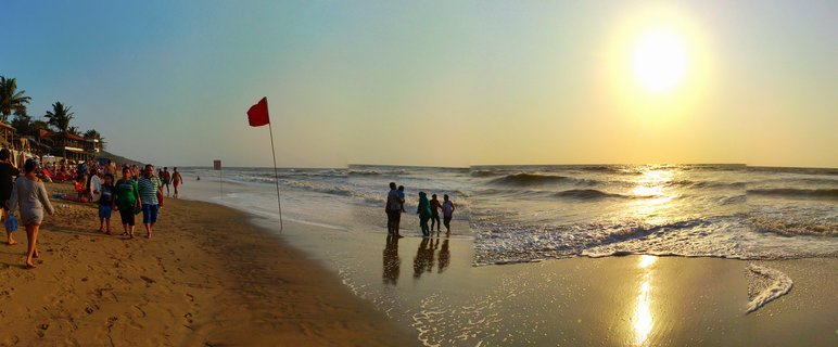 People walking and standing in shallow ocean water on a sandy beach during sunset with palm trees and beach structures visible on the left side