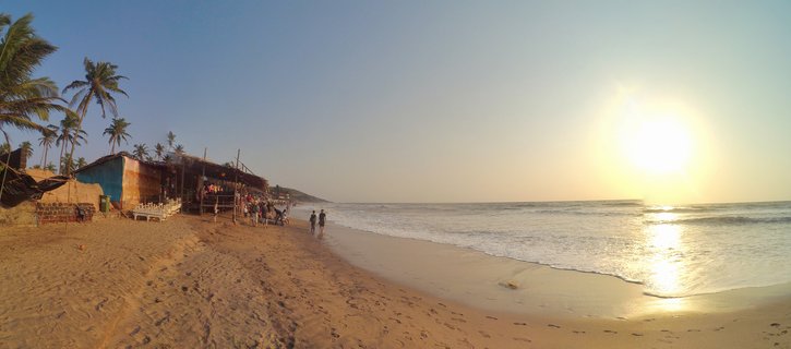 Panoramic beach scene in Goa, India showing palm trees, beach shacks, people walking on sand, and sunset over the ocean