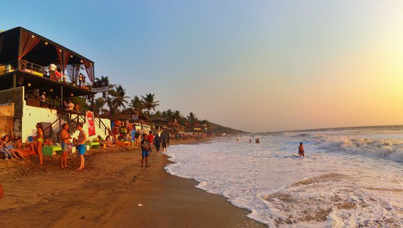 Beach scene at sunset showing people on sand near two-story beach shacks with palm trees, ocean waves rolling onto shore on India's Goa coastline
