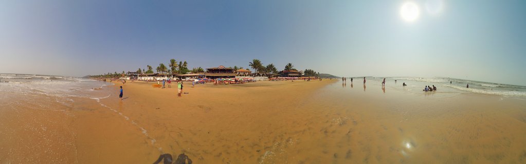 Panoramic beach scene showing golden sand, ocean waves, people walking and swimming, beach shacks with thatched roofs, and palm trees under a clear blue sky with bright sun