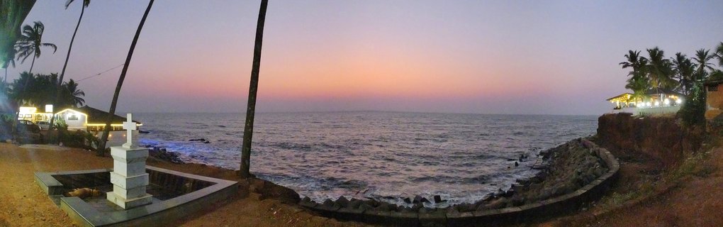 Panoramic beach scene at twilight showing a white cross monument, palm trees, illuminated beach restaurants, and the ocean with a pink-purple sunset sky in Goa, India