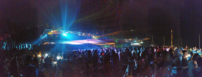 Large crowd of people at outdoor night party in Goa with blue stage lighting beams and canopy tents