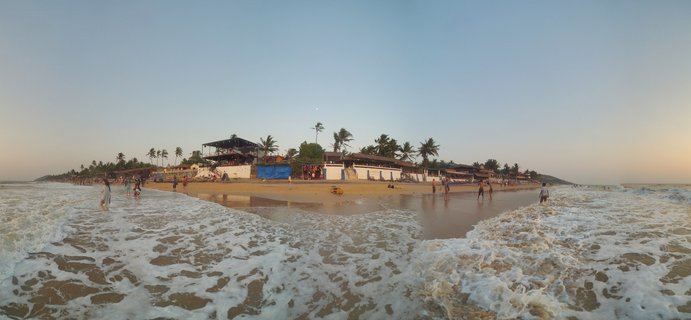 Ocean waves with white foam washing onto a sandy beach in Goa, India, with people walking on the shore, beach buildings and palm trees visible in the background under a clear sky