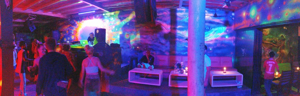 Interior of a nightclub with psychedelic wall murals illuminated by blue and purple neon lights, showing a DJ booth, lounge seating with white tables and candles, and patrons socializing