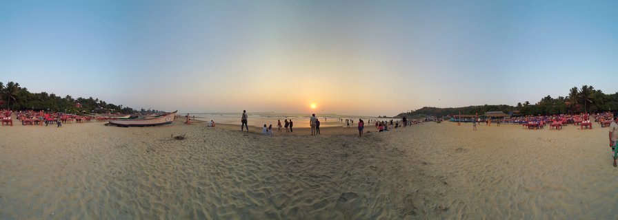Wide panoramic beach scene at sunset in Goa, India showing sand, ocean, a fishing boat, people walking, red beach chairs, palm trees, and golden sun on the horizon