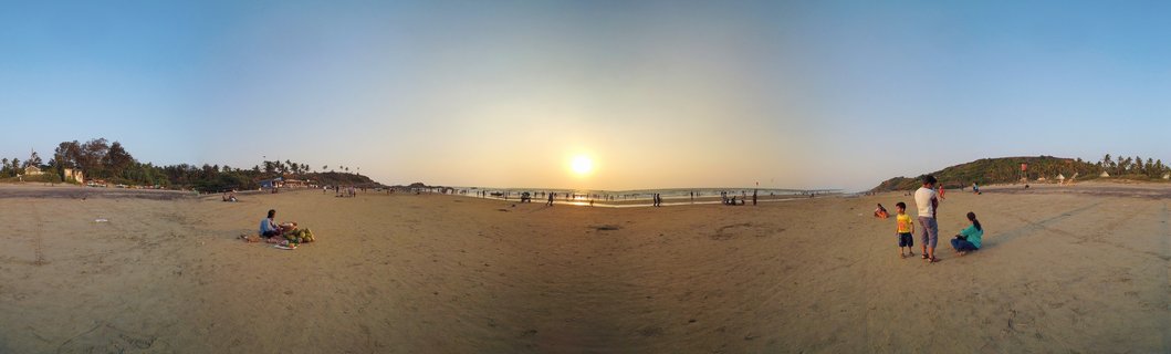 Wide panoramic beach scene at sunset in Goa, India showing sandy shore with multiple groups of people, palm trees on both sides, ocean in the distance, and sun low on the horizon