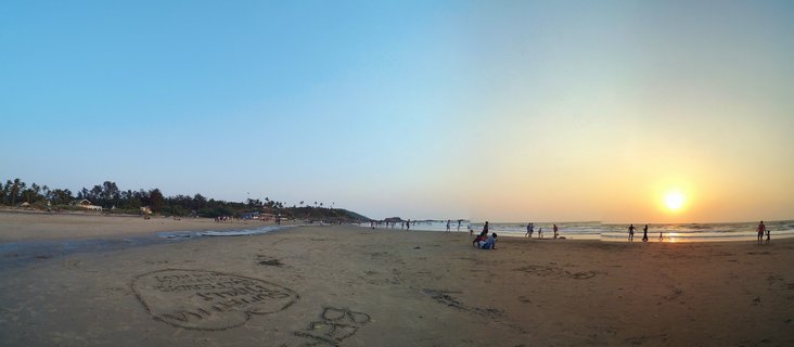Panoramic beach scene at sunset in Goa, India showing silhouettes of people on sand, palm trees on the left, ocean waves on the right, and bright sun low on the horizon with golden sky