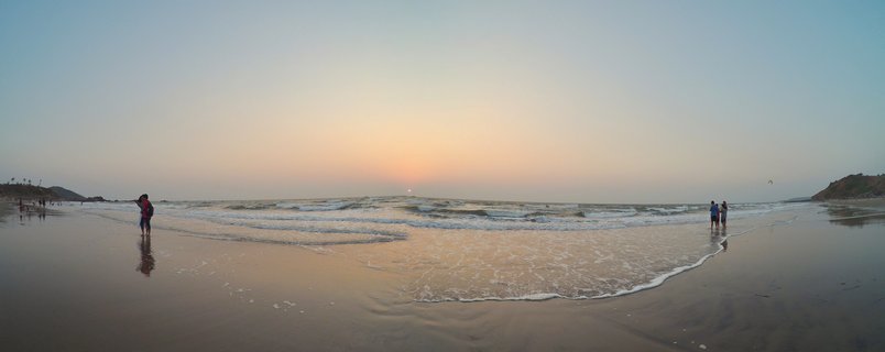 Wide beach panorama at sunset showing ocean waves, wet sand with reflections, silhouettes of people walking, and the sun setting on the horizon with hills visible on both sides